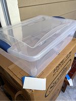 Clear Sterilite plastic storage box with blue latch lid on top of the cardboard box, showing transparency and latch mechanism.