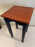 Full view of the side table showing the reddish brown wood top and black painted legs, placed against a neutral wall and white floor.