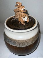 Side view of stoneware jar with glazed brown and cream body, showing animal figurines on the lid