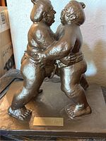 Close-up frontal image of the bronze-toned sumo wrestling plaster cast sculpture on a rectangular base, showing the wrestlers in an embrace with a small plaque labeled 'Sumo Wrestling.'
