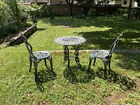 Outdoor photo showing two dark green metal chairs and one round table with ornate rose flower pattern in a grassy yard.