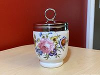 Egg coddler showing floral painted porcelain body with metal lid and loop handle.