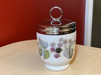 White ceramic egg coddler decorated with blackberry and floral design, metal lid with loop handle on top