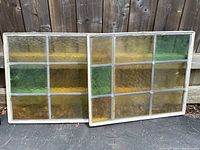 Pair of rectangular leaded stained glass windows resting against wooden fence outdoors, showing full front view of amber, yellow, green panes within leaded grid frames.