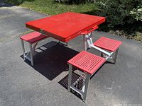 Front-side view of the red portable picnic table with attached seats, showing the table and three seats unfolded and set up outdoors on a paved surface.
