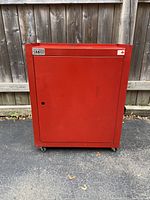 Front view of a red metal tool cabinet with a single door, resting on casters, positioned outdoors on asphalt in front of a wooden fence.