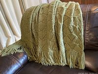 Soft green blanket folded and draped on a dark brown leather couch showing fringe tassels.
