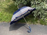 Open umbrella parasol with blue and orange checkered pattern and blue ruffle trim on edge, lying on paved area next to grass and shrub