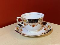 Teacup and saucer displayed showing ornate floral and gold design on white porcelain against a red background.