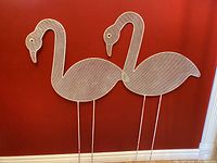 Photo of two vintage flamingo garden picks against a red wall, showing full front and side profile of metal mesh flamingos with metal rod stakes.