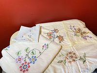 Four vintage embroidered white linen cloths laid on a surface showing detailed colorful embroidery with birds, flowers, and butterflies.