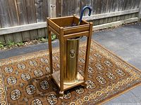Umbrella stand on patterned rug showing wooden frame and brass canister with umbrella inside.