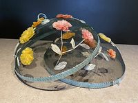 Two vintage wire mesh food covers decorated with colorful raffia flowers on a flat surface against a dark background