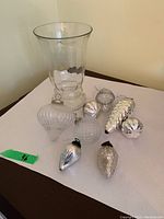 Photo showing eight varying shaped glass Christmas tree ornaments and a clear glass vase on a white surface.