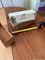 Full view of the vintage Elastoplast metal dispenser showing wear and original box next to it, with measuring tape for scale.