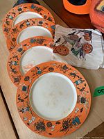 Four vintage Halloween paper plates with orange edges and black cat/witch motifs plus one Halloween paper napkin with witch and pumpkin design on wood surface.