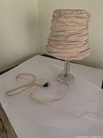 Full view of the small bedside lamp showing beige wrinkled fabric shade, clear glass textured base, and attached power cord with plug