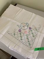 White tablecloth with embroidered teapot, floral accents, and patterned cloth design in green and purple thread.