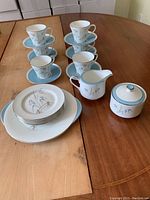 Photo showing 6 cups stacked on matching saucers, 5 luncheon plates, 1 serving plate, milk jug, and sugar bowl arranged on a wooden table.