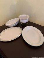 Photo of two oval white platters with green trim, three smaller oval bakeware dishes, and a white ceramic chocolate pot on metal stand taken on brown table against beige wall.