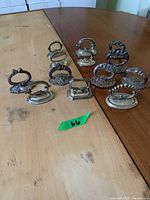 Photo of 13 varying silver handle napkin rings on a wooden table, showing different designs including smooth and ornate bases with loop and rounded handles.