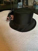 Front view of black top hat showing smooth fabric finish, wide ribbon band, and attached feather and small pink flower adornment.