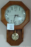 Front view of wooden wall clock with visible octagonal frame, white clock face with black numbers, brass-colored pendulum below, and seller's lot tag attached.