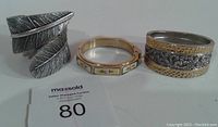 Front view of three bracelets showing the feather clamp style silver-tone bracelet, octagon rose tile bangle with floral designs, and the wide two-toned hammered and filigree bangle