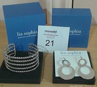 Two blue Lia Sophia jewelry boxes behind a silver-tone multi-band cuff bracelet and a pair of double circle earrings on a black foam base with a Lia Sophia display card and auction sticker.