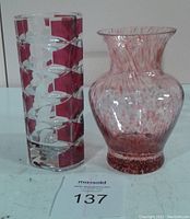 Two vases placed side by side on a flat surface, one tall clear glass with red horizontal bands, one shorter red and white speckled glass vase