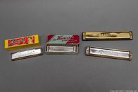 Four vintage harmonicas laid out with their original boxes on a gray background, showing condition and style.