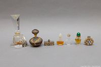 Front view of seven assorted vintage perfume bottles in a row, showing various shapes, colors, and stoppers including crystal, metallic, ornate gold-tone, and decorative items.
