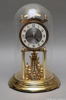 Full view of the domed mantle clock showing brass base, rotating pendulum balls, clear glass dome, clock face with metal and white contrast dial.