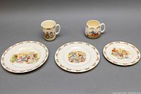 Full view of two Royal Doulton Bunnykin mugs and three Bunnykin plates of sizes 8.5 inch, 8 inch, and 6.5 inch on display surface.