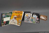 Overview of two vintage postage stamp albums, loose stamps in plastic cases, and a small box of stamps arranged on a grey surface.