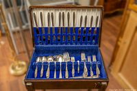 Opened wood chest stand with blue velvet lining, displaying various silver plated flatware pieces in arranged slots.