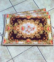 Three identical rectangular area rugs stacked on tiled floor, showing floral and ornate design with pink roses and gold ornamental border.