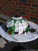 Photo showing dome-shaped stained glass lamp shade with white glass panels, green leaves, and grape clusters in purple.