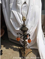 Full view of tall brass Hollywood Regency lamp with three orange lucite dangling ornaments, bulb socket with harp and no shade, placed against white draped background