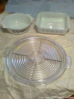 Round clear glass Manhattan platter with concentric circle design and handles alongside two Pyrex dishes: one square with stylized floral pattern, one round with brown floral pattern.