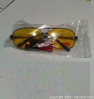 Clear photo of yellow tinted night driving glasses in sealed clear plastic packaging, showing metal frame and black temple tips.