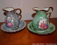 Photo showing two ceramic pitchers on matching plates with gold trim, one blue, one green, decorated with vintage figure scenes