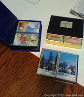 Photo of six decks of playing cards: two with animal artwork in blue cases, two in a black Congress Playing Cards box, two with mountain landscape backs in plastic case.