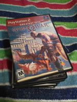Stack of PlayStation 2 video game cases with God of War Greatest Hits on top, showing cover art of Kratos fighting a creature.