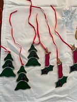 Photo shows stained glass Christmas ornaments including three green trees and three red candles with holly, arranged on white cloth with red ribbon hangers visible.