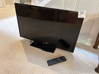Hitachi 29" LCD TV with remote control on carpeted floor, showing front angle view with reflection visible on screen.