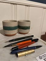 Photo showing two speckled beige ceramic pottery cups with green glaze band and five writing instruments including mechanical pencils and a calligraphy pen.