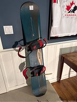 Santa Cruz SC snowboard standing upright against a blue wall with attached black and red bindings, sunlight highlighting surface and showing wear.