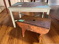 Side table with peeling paint on top surface, decorative carved border, cross legs, and small antique pine stool with hand hole and aged wood patina.