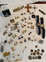 Wide shot of cuff links, tie clips, pins, watches, and coins arranged on a white cloth showing various items including brass fish tie clip and Fossil watches